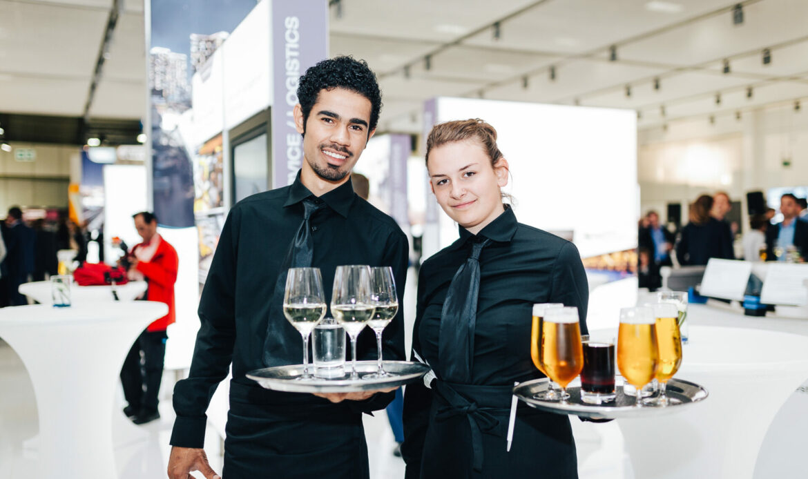 Zwei Kellner*innen mit vollen Tabletts bei der Arbeit auf einer Messe